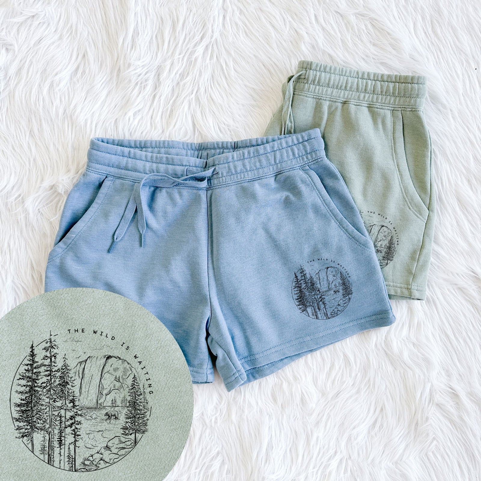 Two pairs of Cali Wave shorts in different colors printed with a round black ink scene of a waterfall, pine trees and a river, with the words The Wild Is Waiting, with a close-up of the print.