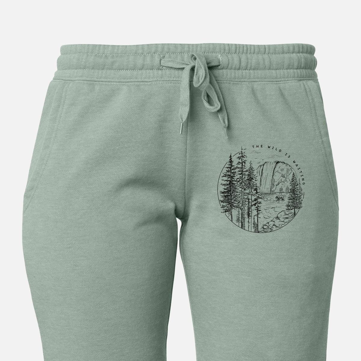 Front of sage green Cali Wave sweatpants, printed with a round black ink scene of a waterfall, pine trees and a river, with the words The Wild Is Waiting on the leg.