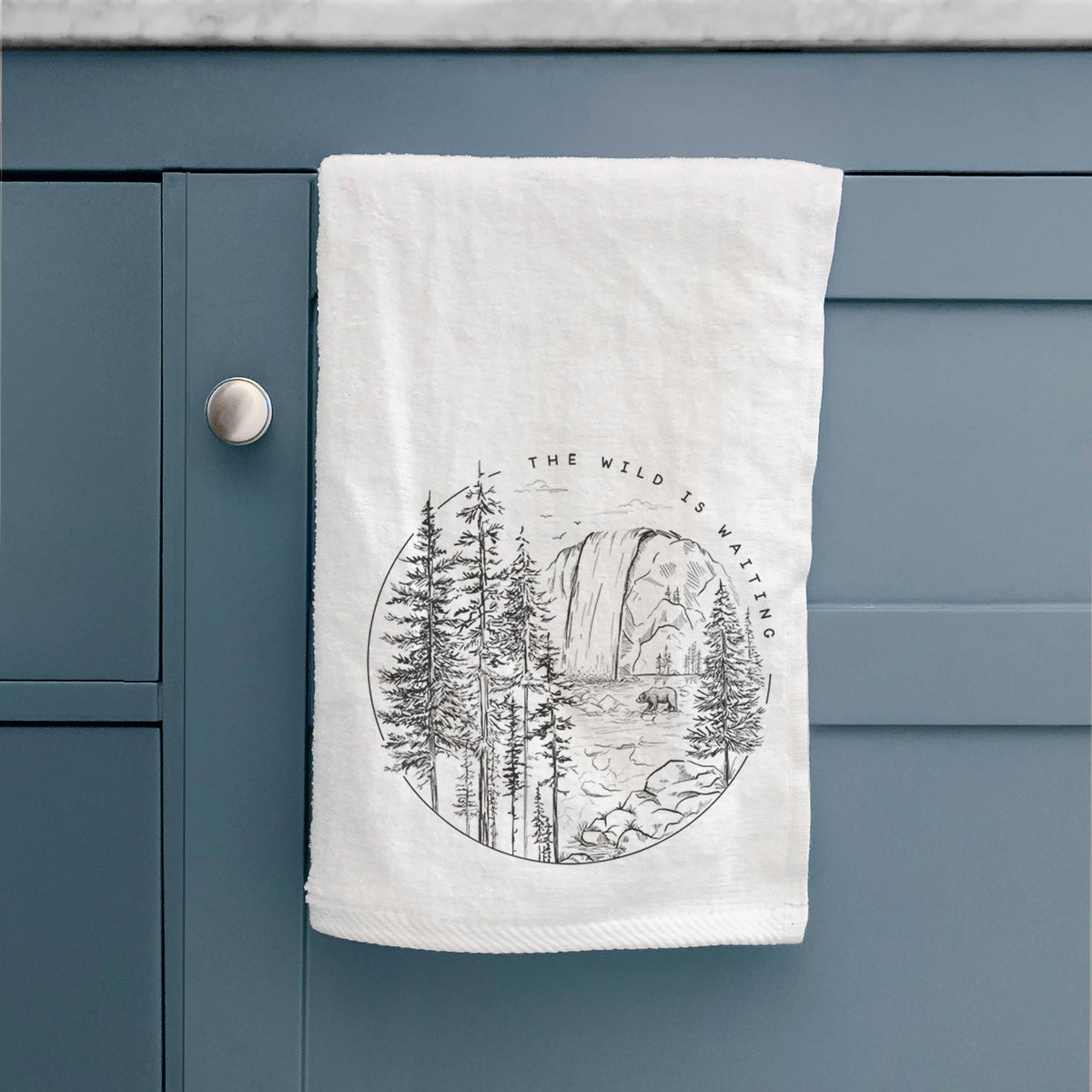 White hand towel printed with a round black ink scene of a waterfall, pine trees and a river, with the words The Wild Is Waiting, hanging on a cabinet door.