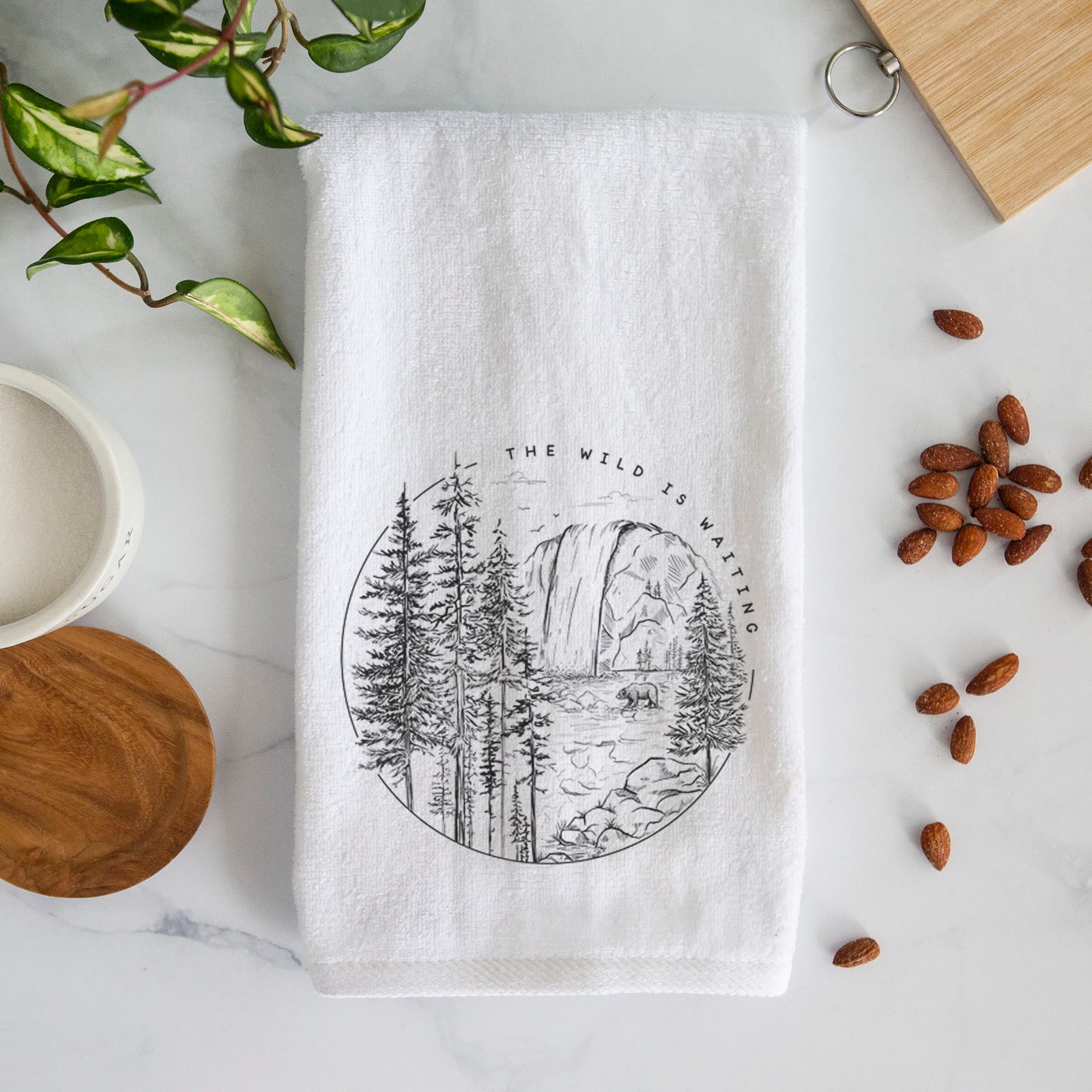 White hand towel printed with a round black ink scene of a waterfall, pine trees and a river, with the words The Wild Is Waiting.