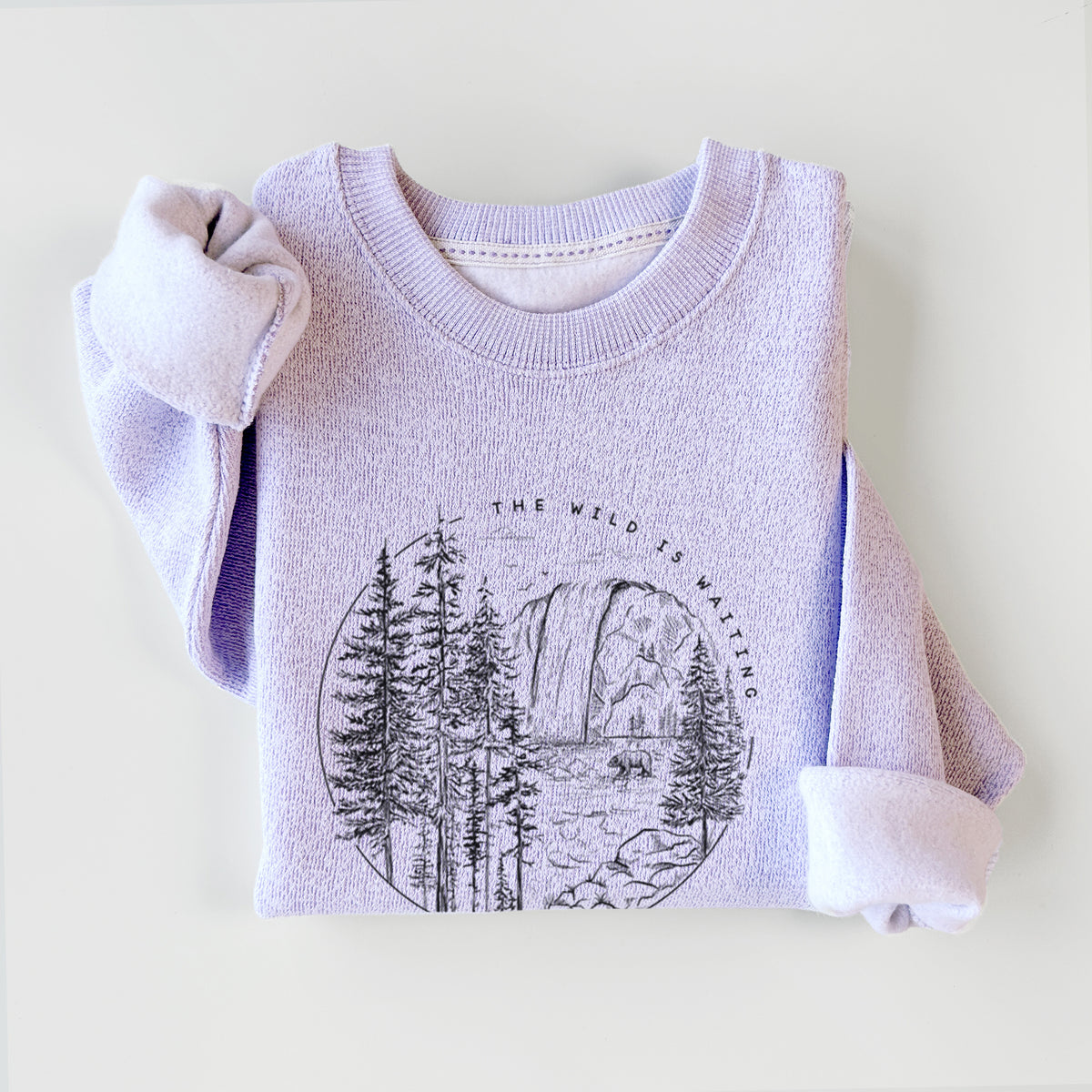 Ice purple knit sweatshirt printed with a round black ink scene of a waterfall, pine trees and a river, with the words The Wild Is Waiting, styled flat.