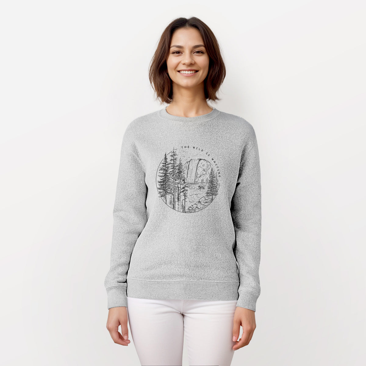 Woman wearing a knit sweatshirt printed with a round black ink scene of a waterfall, pine trees and a river, with the words The Wild Is Waiting.