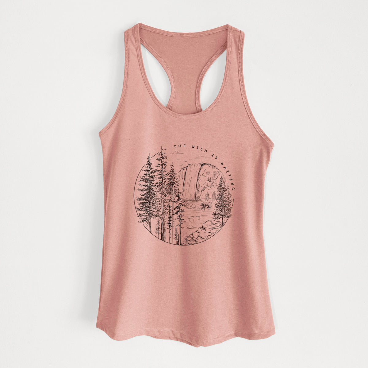 Desert pink women's racerback tank top laid flat, printed with a round black ink scene of a waterfall, pine trees and a river, with the words The Wild Is Waiting.