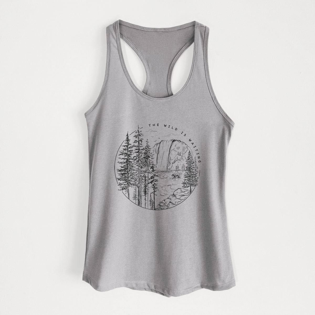 Heather grey women's racerback tank top laid flat, printed with a round black ink scene of a waterfall, pine trees and a river, with the words The Wild Is Waiting.