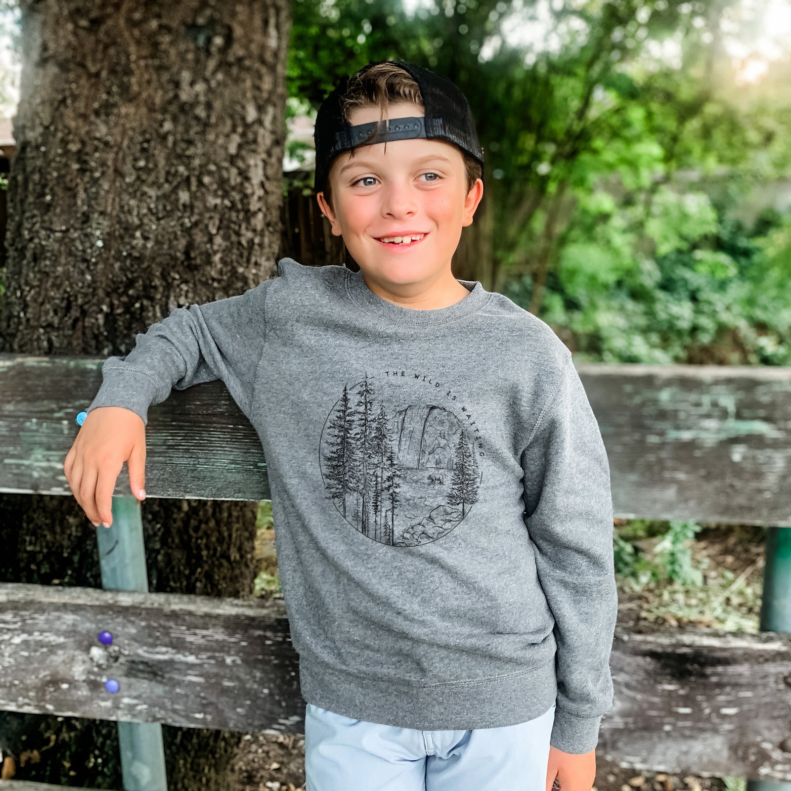 Boy wearing a youth crewneck sweatshirt printed with a round black ink scene of a waterfall, pine trees and a river, with the words The Wild Is Waiting, outdoors.