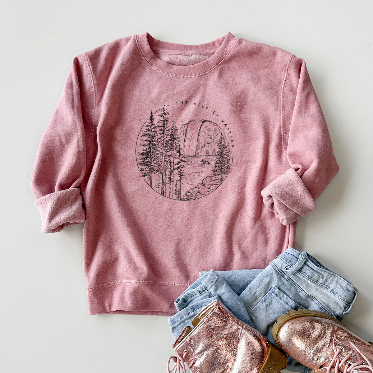 Youth crewneck sweatshirt printed with a round black ink scene of a waterfall, pine trees and a river, with the words The Wild Is Waiting, styled flat with jeans and boots.