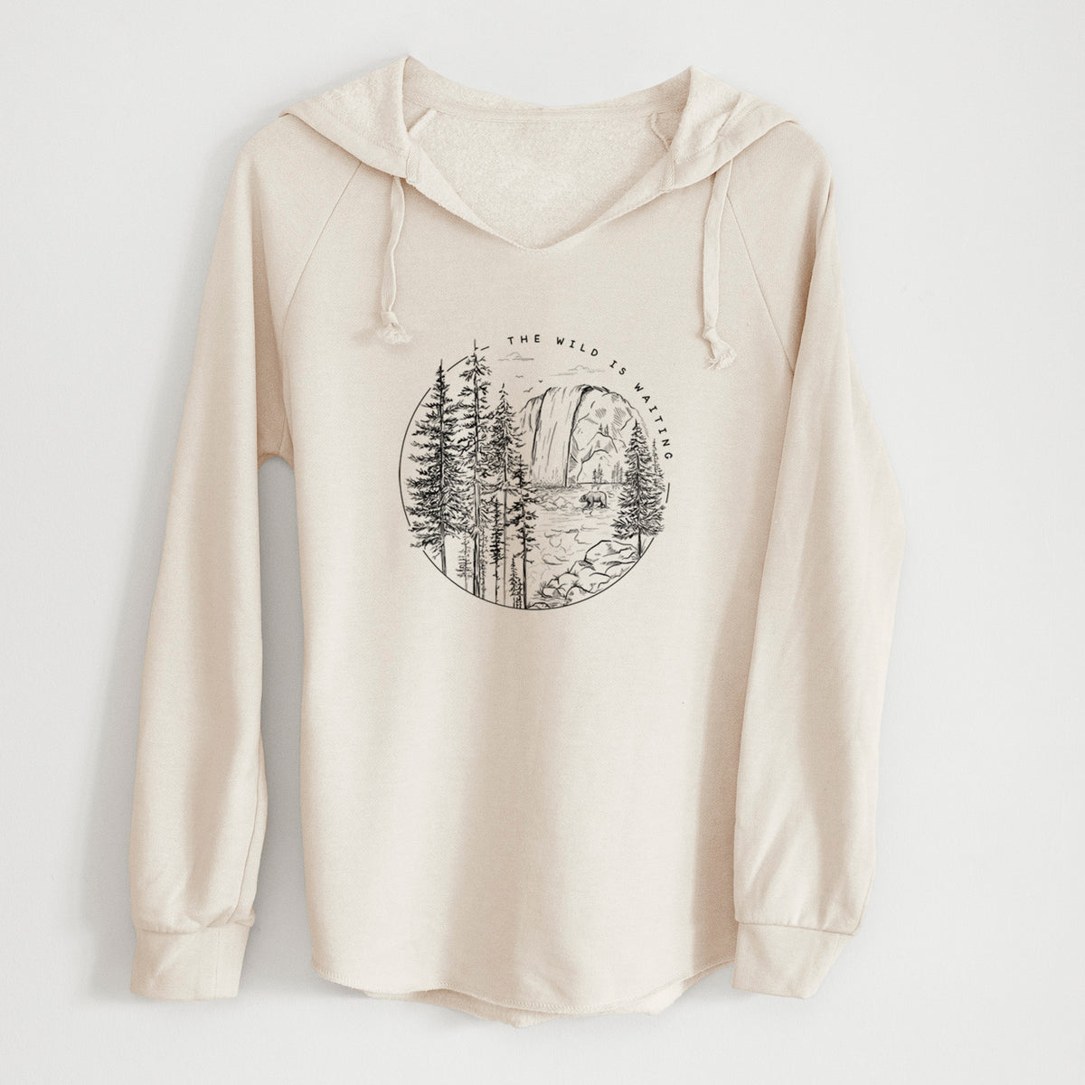 Bone cream women's Cali Wave hooded sweatshirt laid flat, printed with a round black ink scene of a waterfall, pine trees and a river, with the words The Wild Is Waiting.