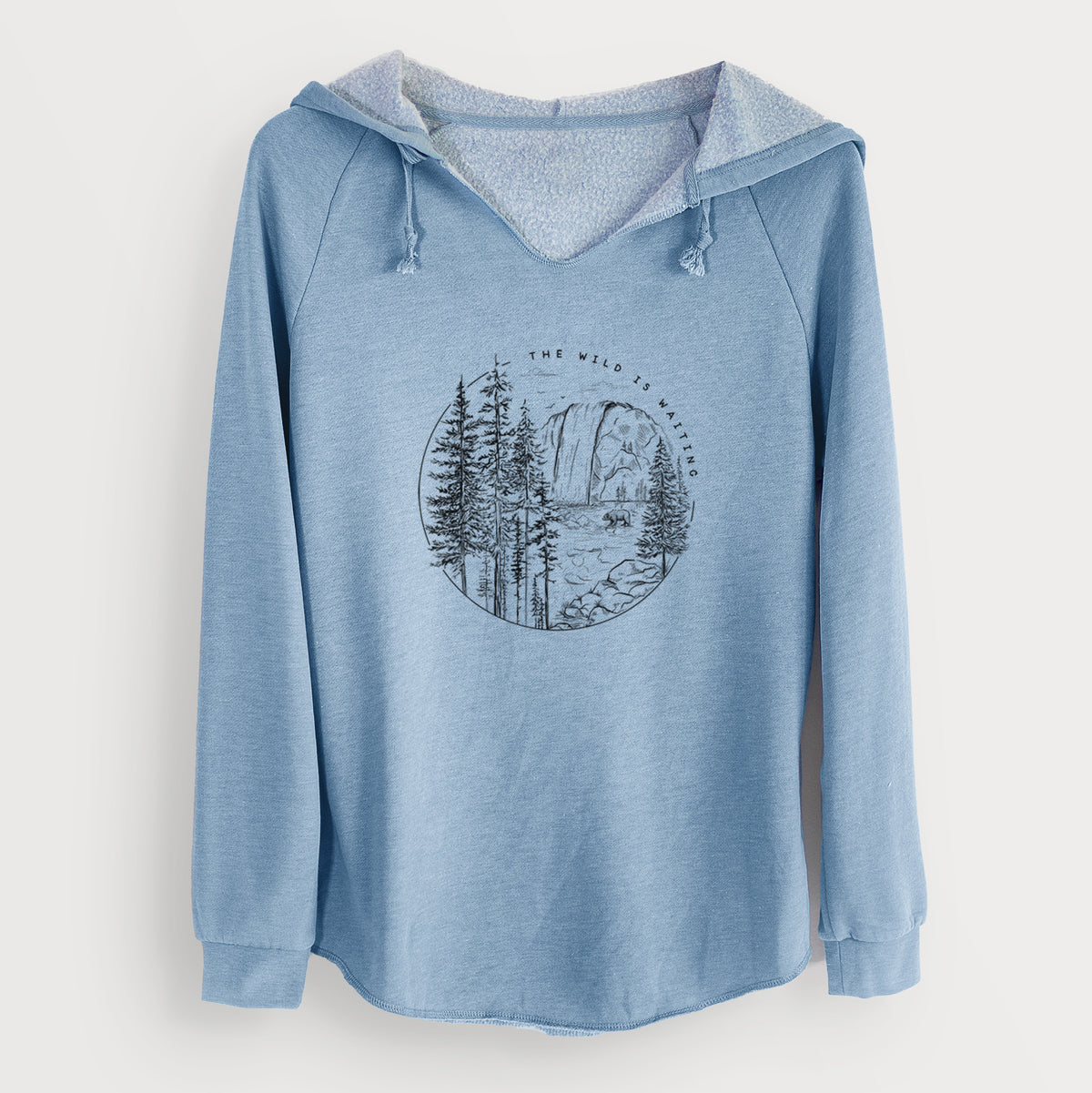 Misty blue women's Cali Wave hooded sweatshirt laid flat, printed with a round black ink scene of a waterfall, pine trees and a river, with the words The Wild Is Waiting.