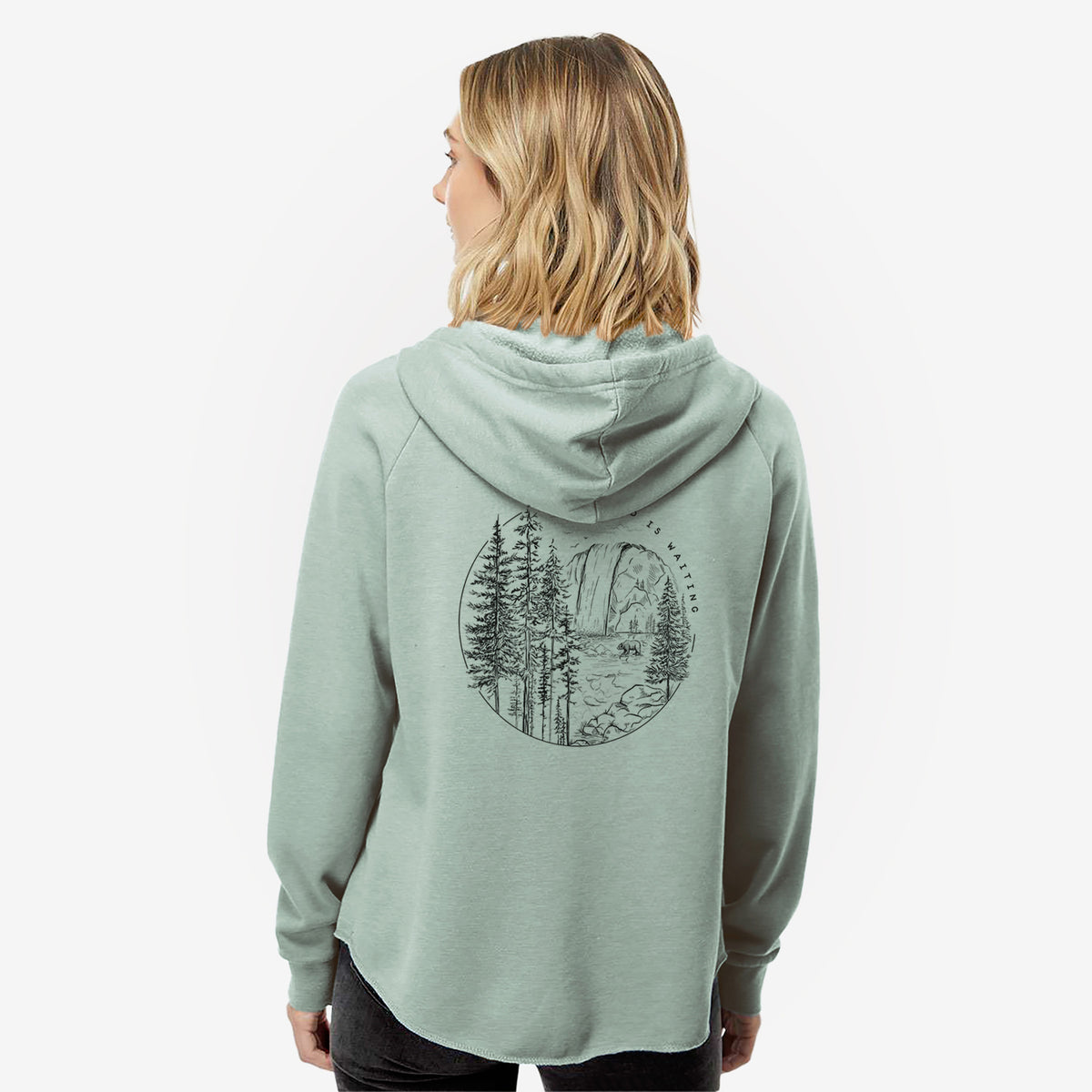 Woman wearing a sage green women's Cali Wave zip-up sweatshirt printed with a round black ink scene of a waterfall, pine trees and a river, with the words The Wild Is Waiting, seen from behind.