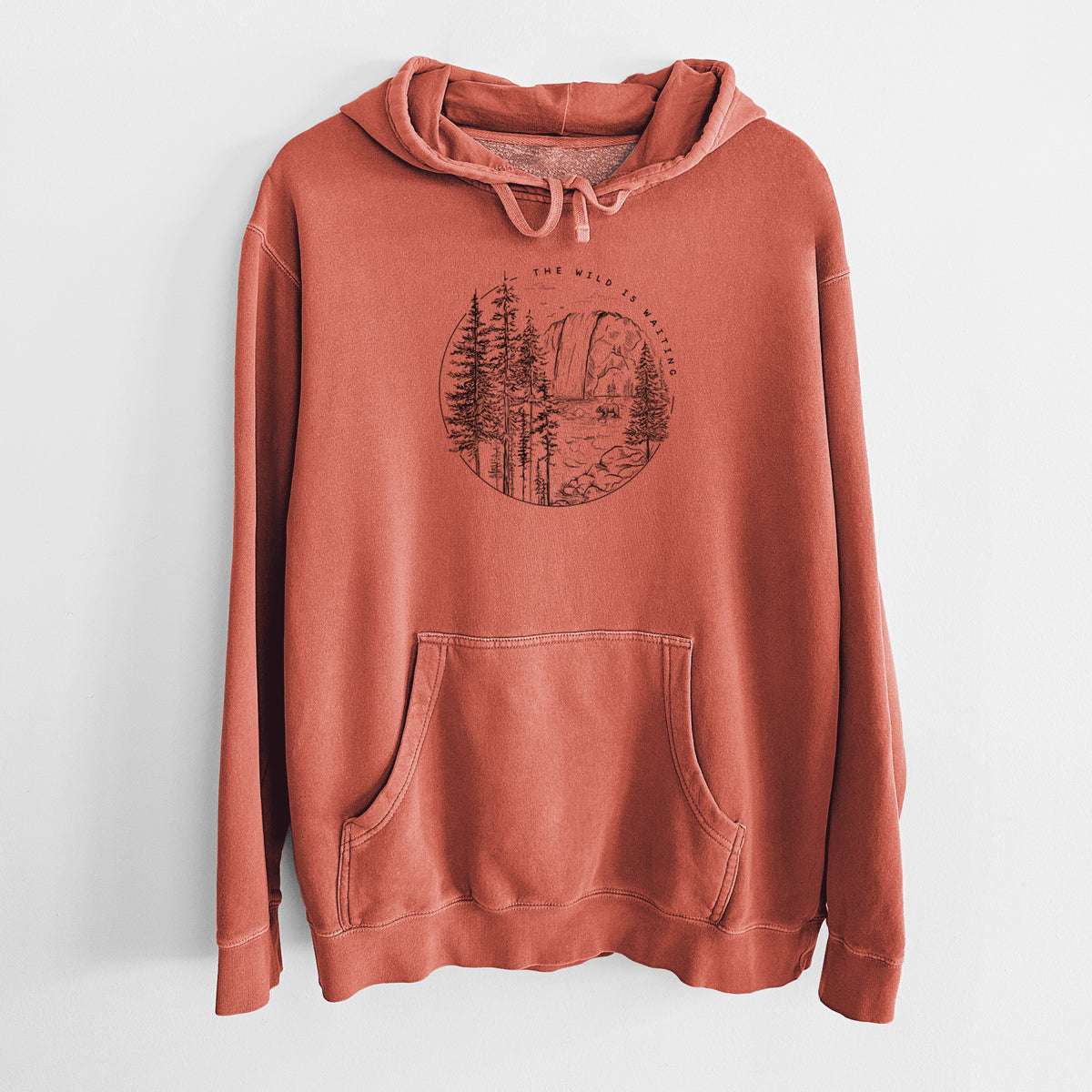 Amber orange pigment dyed hoodie laid flat, printed with a round black ink scene of a waterfall, pine trees and a river, with the words The Wild Is Waiting.