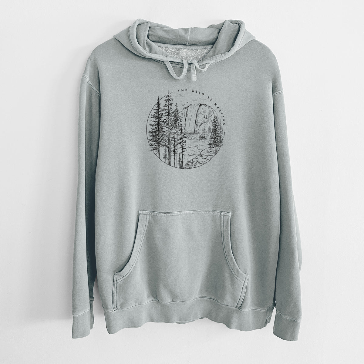 Sage green pigment dyed hoodie laid flat, printed with a round black ink scene of a waterfall, pine trees and a river, with the words The Wild Is Waiting.