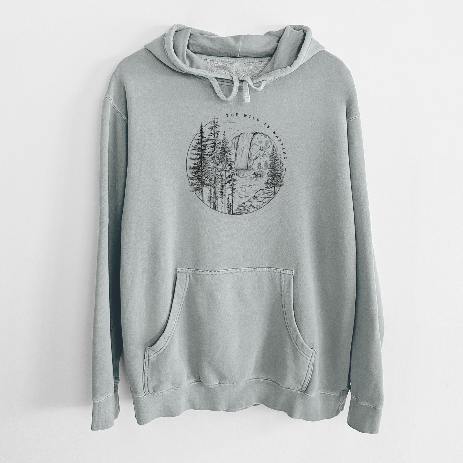 Sage green pigment dyed hoodie laid flat, printed with a round black ink scene of a waterfall, pine trees and a river, with the words The Wild Is Waiting.