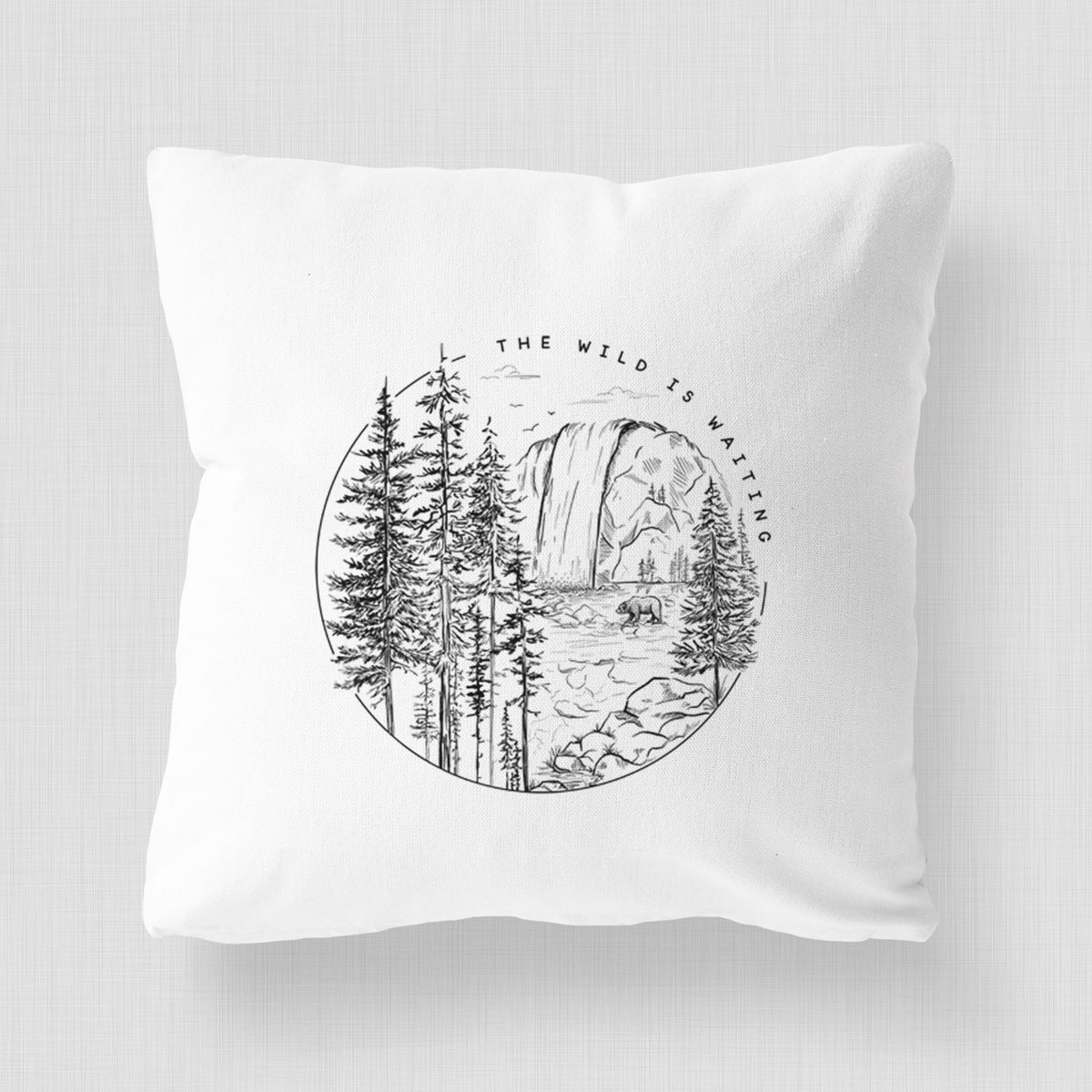 White pillow cover printed with a round black ink scene of a waterfall, pine trees and a river, with the words The Wild Is Waiting.
