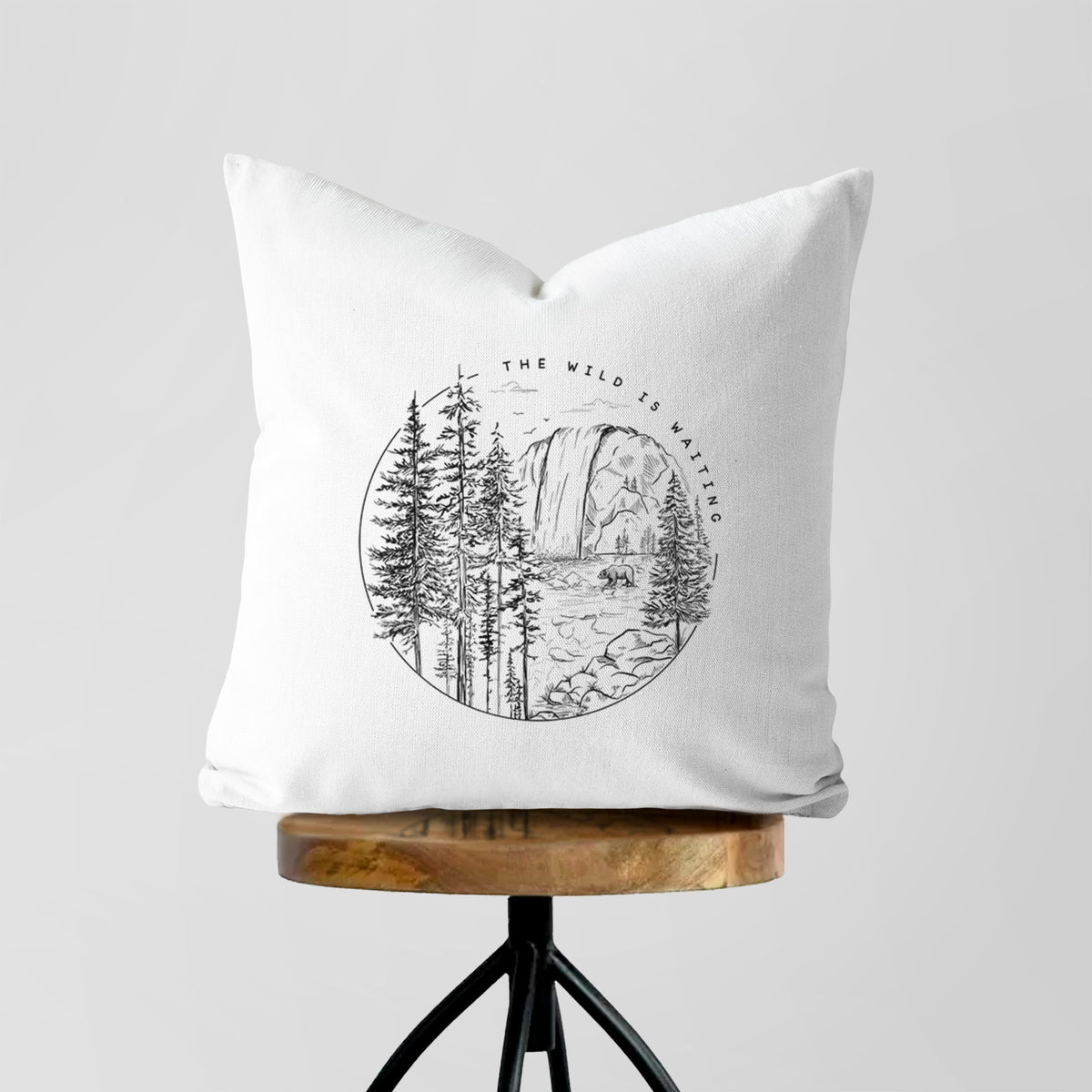 White pillow cover printed with a round black ink scene of a waterfall, pine trees and a river, with the words The Wild Is Waiting, on a wooden stool.