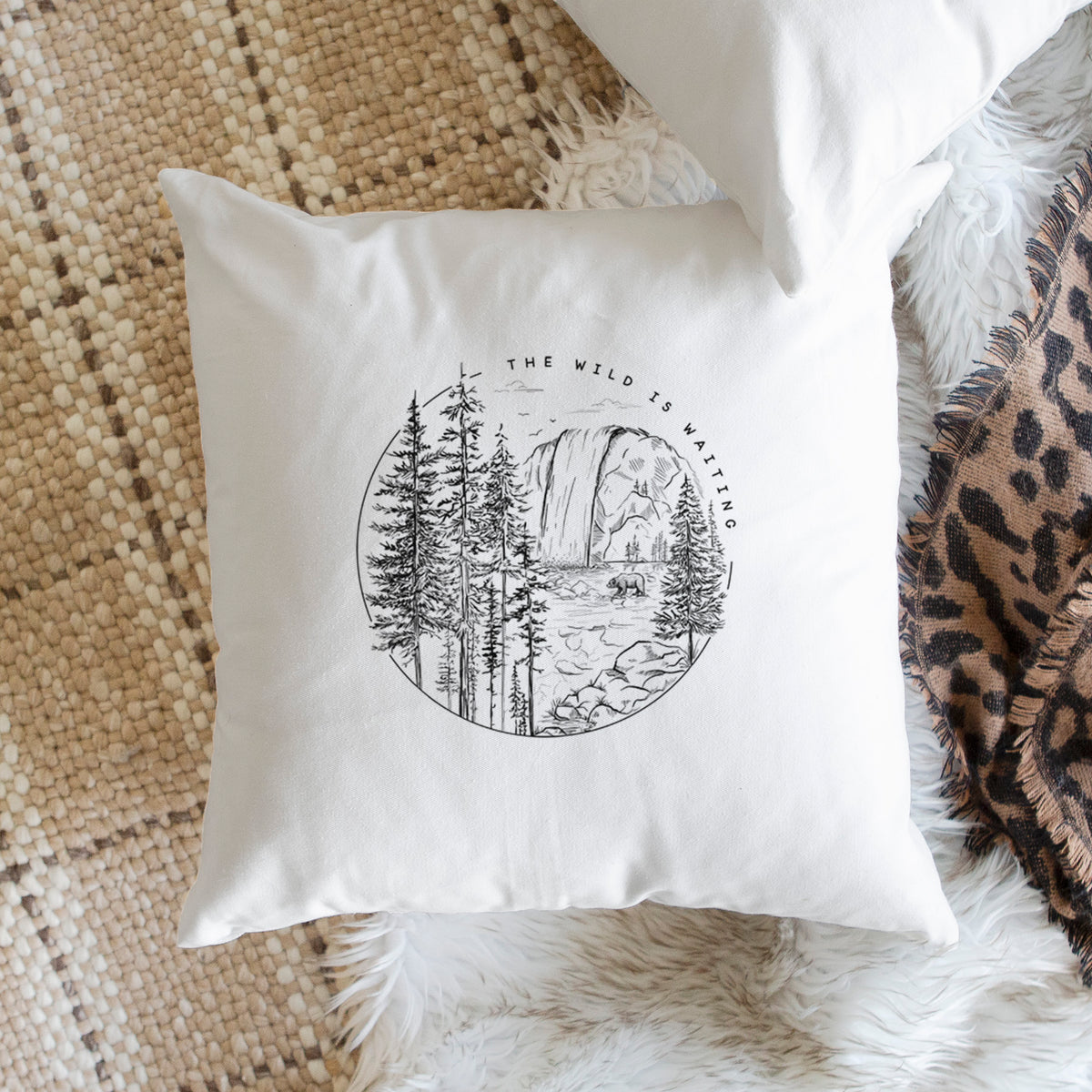White pillow cover printed with a round black ink scene of a waterfall, pine trees and a river, with the words The Wild Is Waiting, on a woven chair.