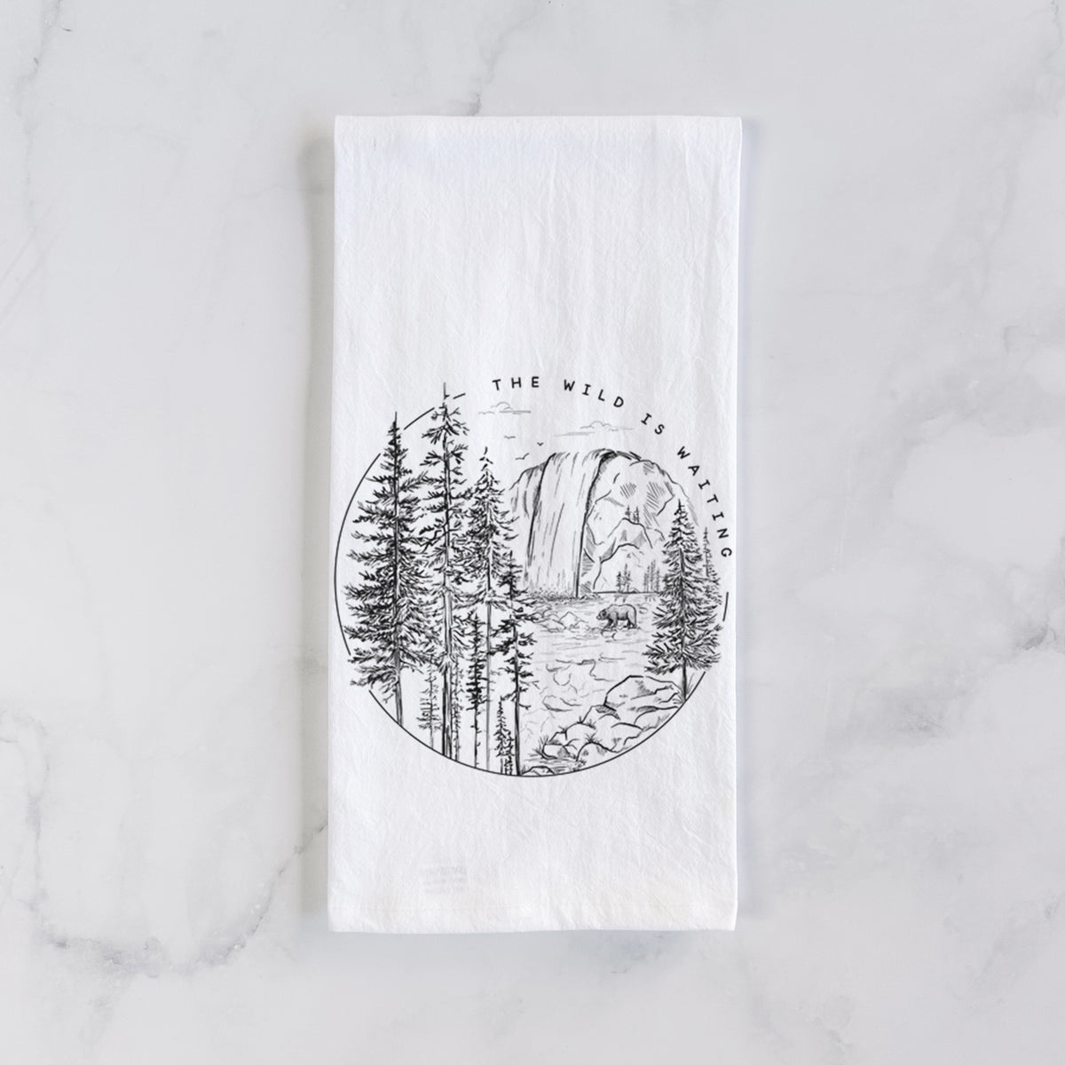 White cotton tea towel laid flat on marble, printed with a round black ink scene of a waterfall, pine trees and a river, with the words The Wild Is Waiting.