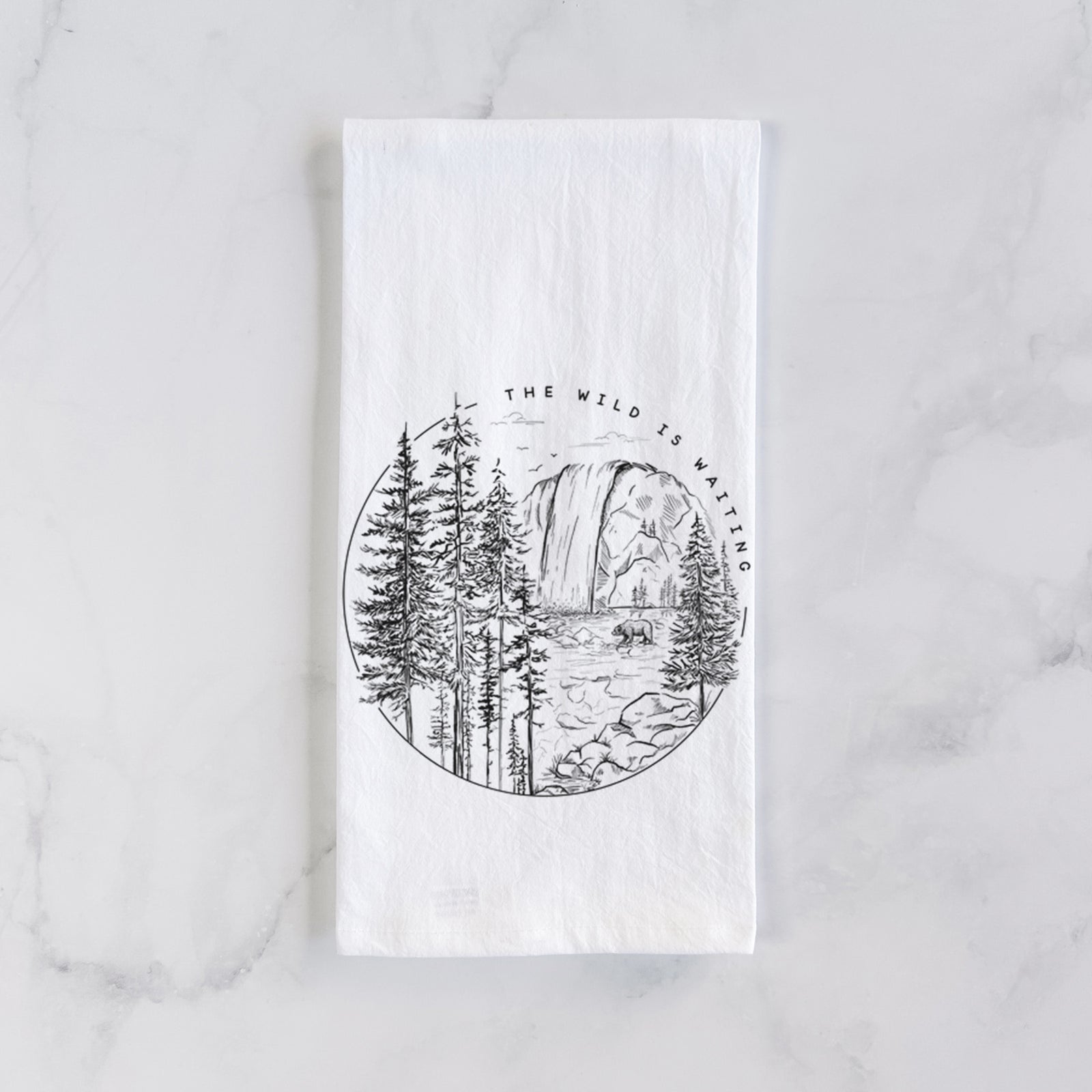 White cotton tea towel printed with a round black ink scene of a waterfall, pine trees and a river, with the words The Wild Is Waiting, on a marble counter beside a round wooden cutting board and greenery.