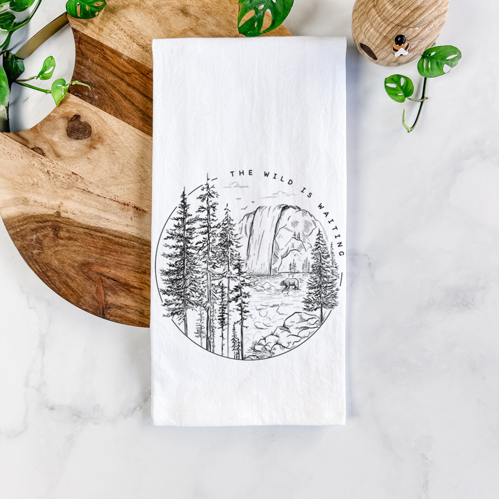 White cotton tea towel printed with a round black ink scene of a waterfall, pine trees and a river, with the words The Wild Is Waiting, on a marble counter beside a round wooden cutting board and greenery.