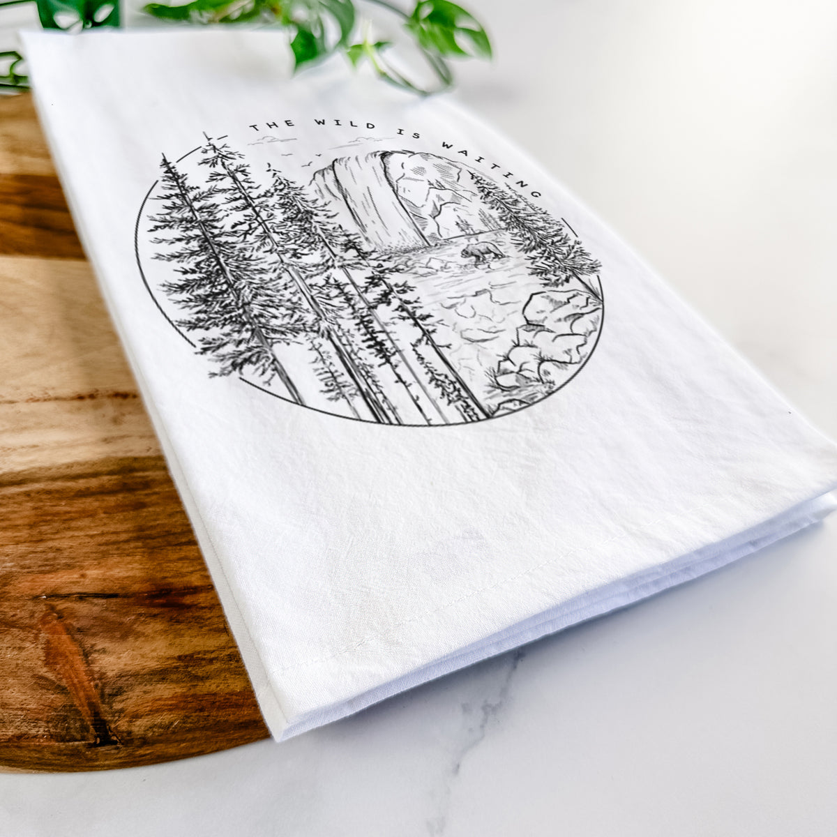 Close-up of a folded white tea towel on a wooden board, showing a round black ink scene of a waterfall, pine trees and a river, with the words The Wild Is Waiting.
