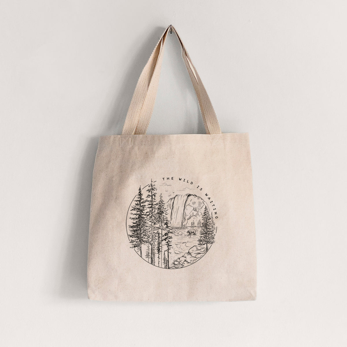 Natural canvas tote bag printed with a round black ink scene of a waterfall, pine trees and a river, with the words The Wild Is Waiting.
