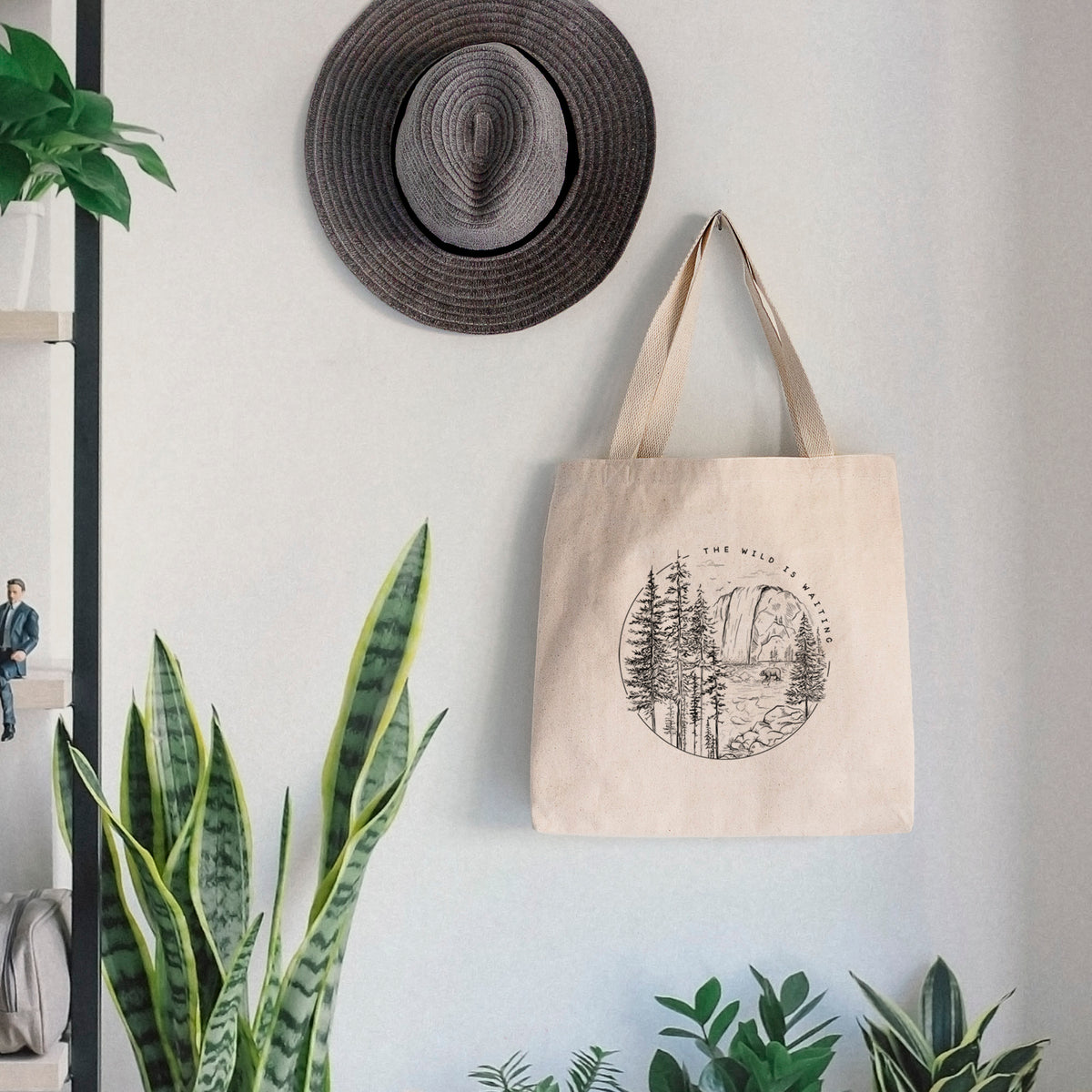 Canvas tote bag printed with a round black ink scene of a waterfall, pine trees and a river, with the words The Wild Is Waiting, hanging on a wall hook below a hat, next to potted plants.