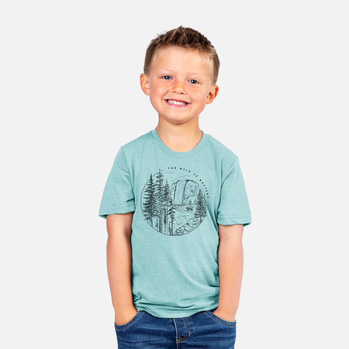 Smiling boy wearing a youth t-shirt printed with a round black ink scene of a waterfall, pine trees and a river, with the words The Wild Is Waiting.