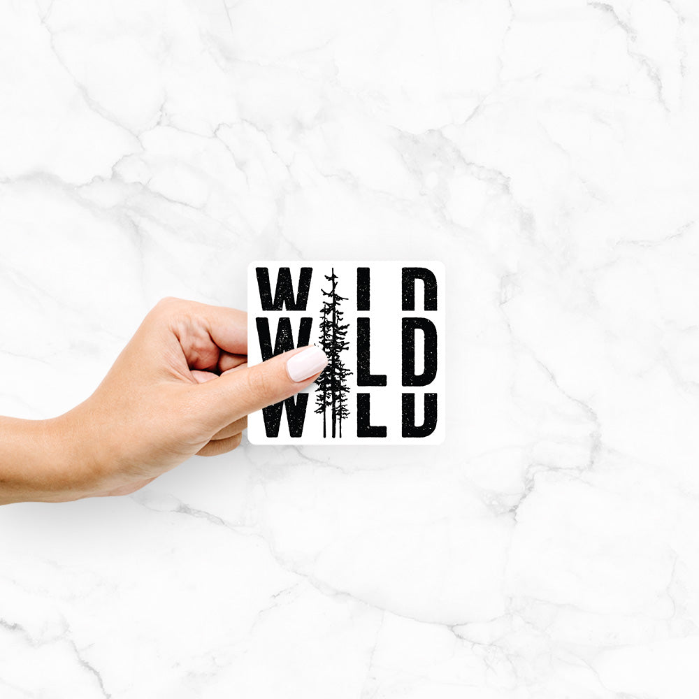 Hand holding a 4 inch vinyl decal of bold black text reading WILD stacked three times, with a tall pine tree forming the letter I.