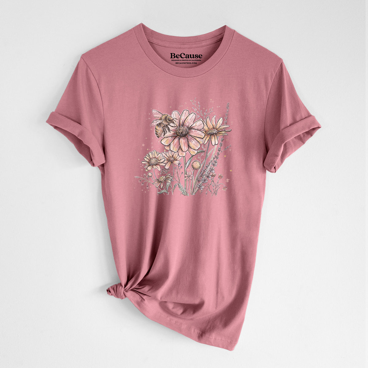 Mauve pink lightweight cotton t-shirt laid flat with the sleeves rolled, printed with a soft full-color illustration of a bee flying over pink and purple wildflowers.