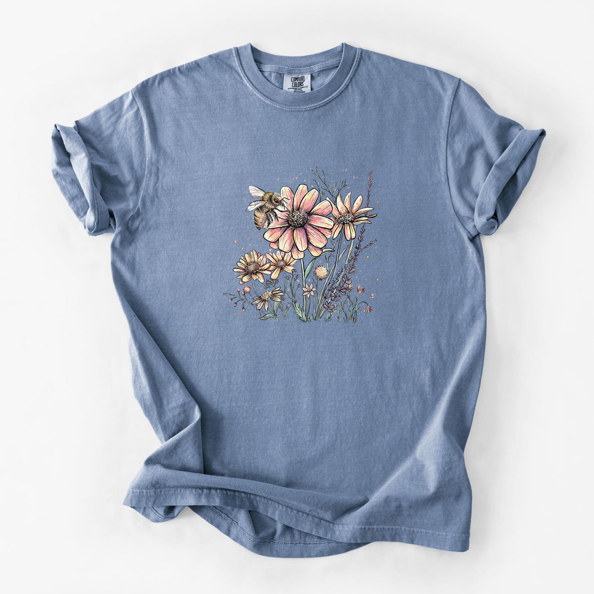 Blue jean Comfort Colors t-shirt laid flat, printed with a soft full-color illustration of a bee flying over pink and purple wildflowers.