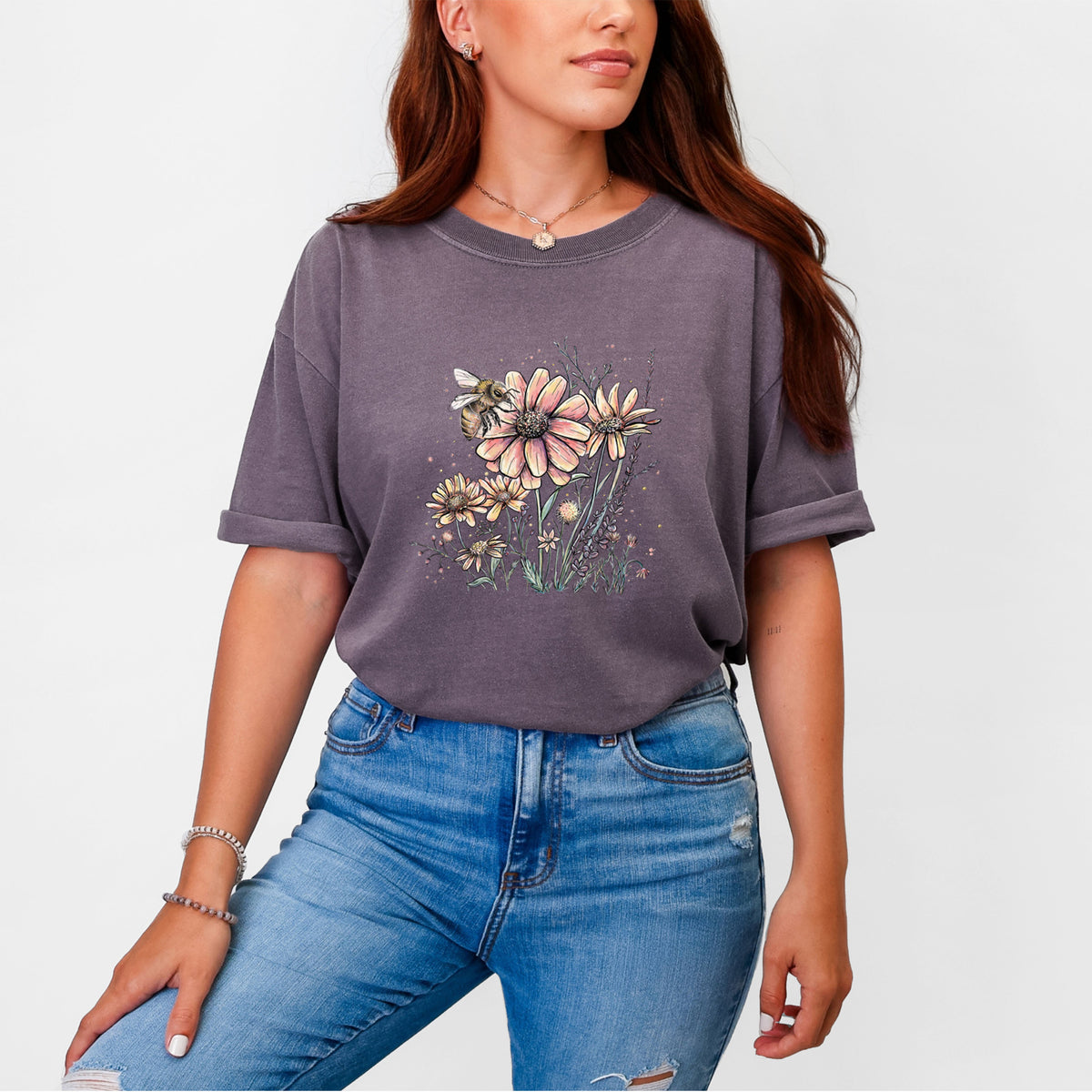 Woman wearing a Comfort Colors t-shirt printed with a soft full-color illustration of a bee flying over pink and purple wildflowers.