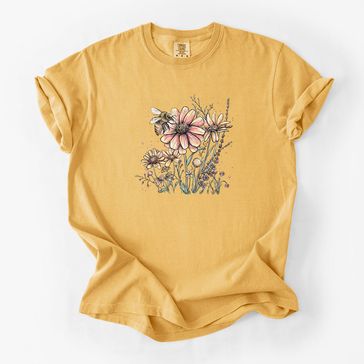Golden wheat yellow Comfort Colors t-shirt laid flat, printed with a soft full-color illustration of a bee flying over pink and purple wildflowers.