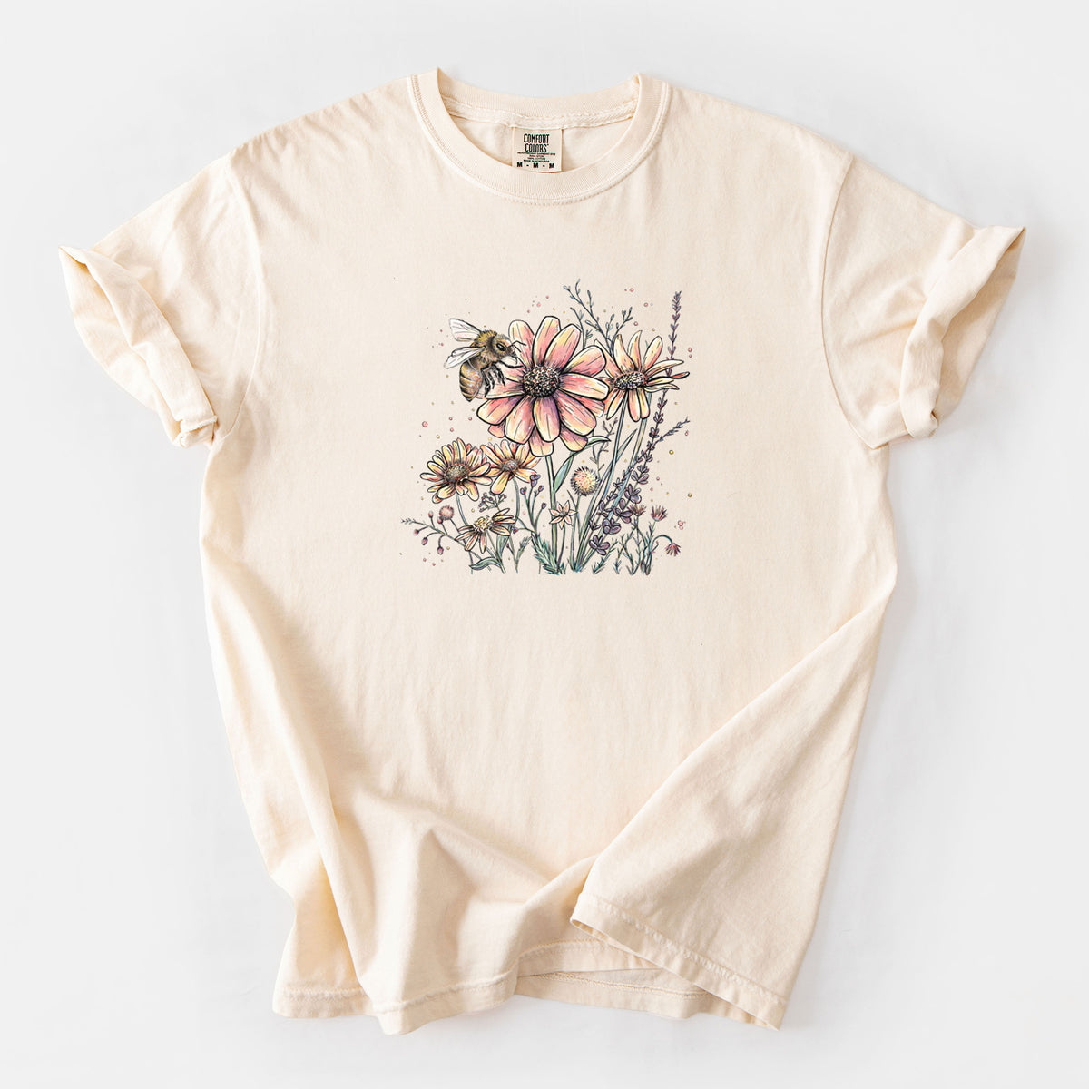 Ivory Comfort Colors t-shirt laid flat, printed with a soft full-color illustration of a bee flying over pink and purple wildflowers.