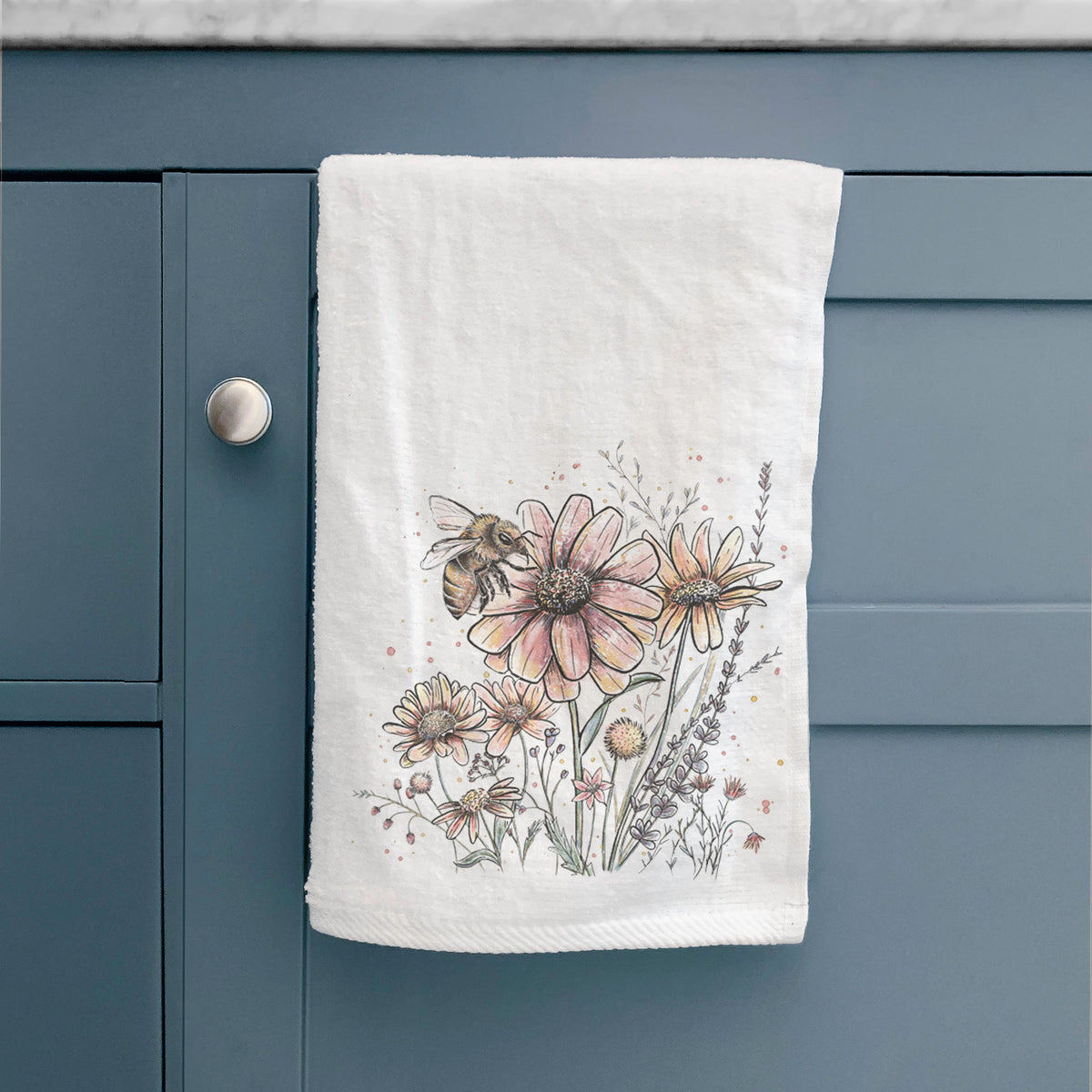 White hand towel printed with a soft full-color illustration of a bee flying over pink and purple wildflowers, hanging on a cabinet door.