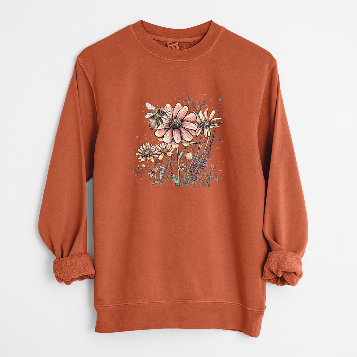Amber orange pigment dyed crewneck sweatshirt laid flat with the sleeves rolled, printed with a soft full-color illustration of a bee flying over pink and purple wildflowers.