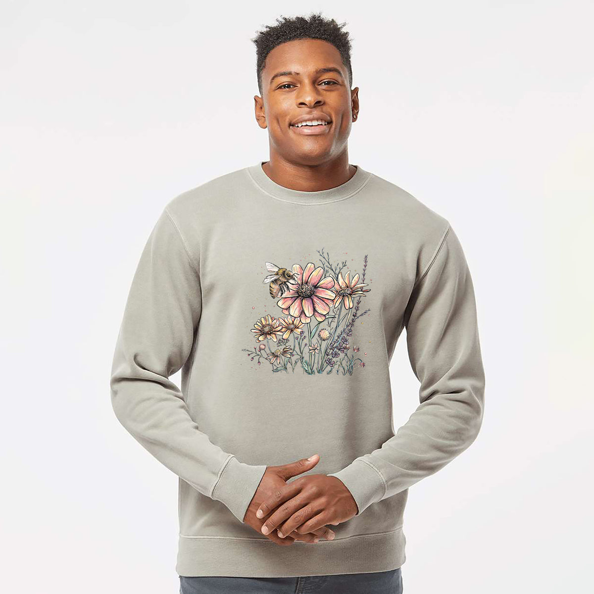 Model wearing a cement grey pigment dyed crewneck sweatshirt printed with a soft full-color illustration of a bee flying over pink and purple wildflowers.