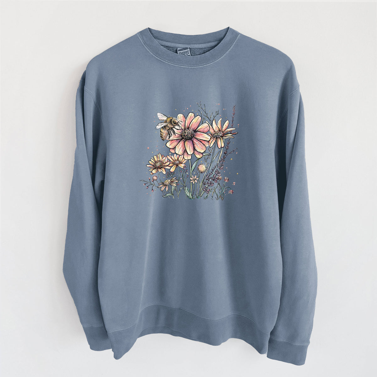 Slate blue pigment dyed crewneck sweatshirt laid flat, printed with a soft full-color illustration of a bee flying over pink and purple wildflowers.