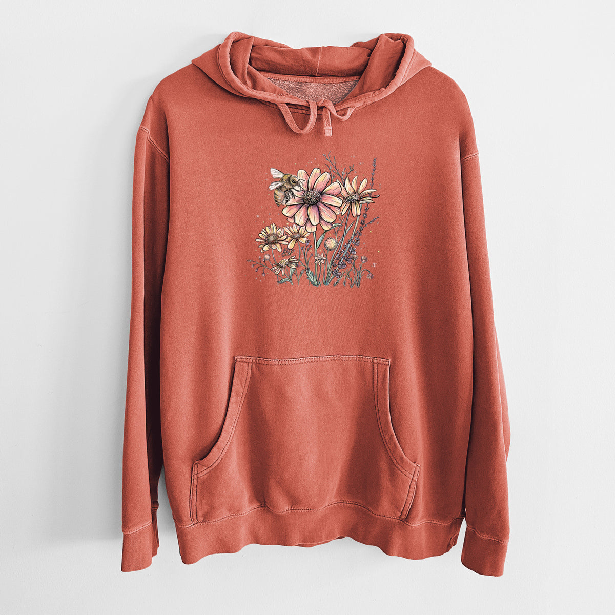Amber orange pigment dyed hoodie laid flat, printed with a soft full-color illustration of a bee flying over pink and purple wildflowers.