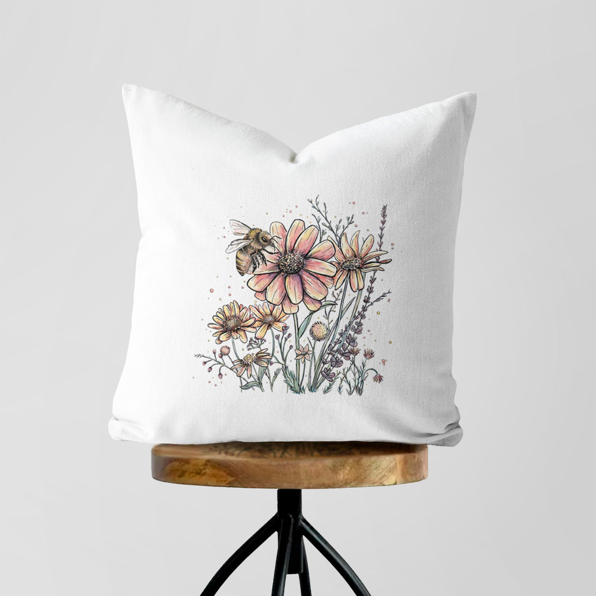 White pillow cover printed with a soft full-color illustration of a bee flying over pink and purple wildflowers, on a wooden stool.