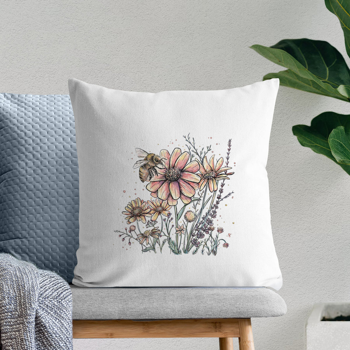 White pillow cover printed with a soft full-color illustration of a bee flying over pink and purple wildflowers, on a bench beside plants.