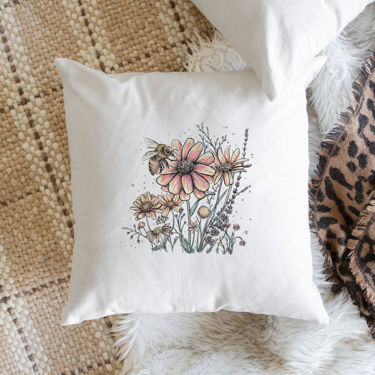 White pillow cover printed with a soft full-color illustration of a bee flying over pink and purple wildflowers, on a woven chair.