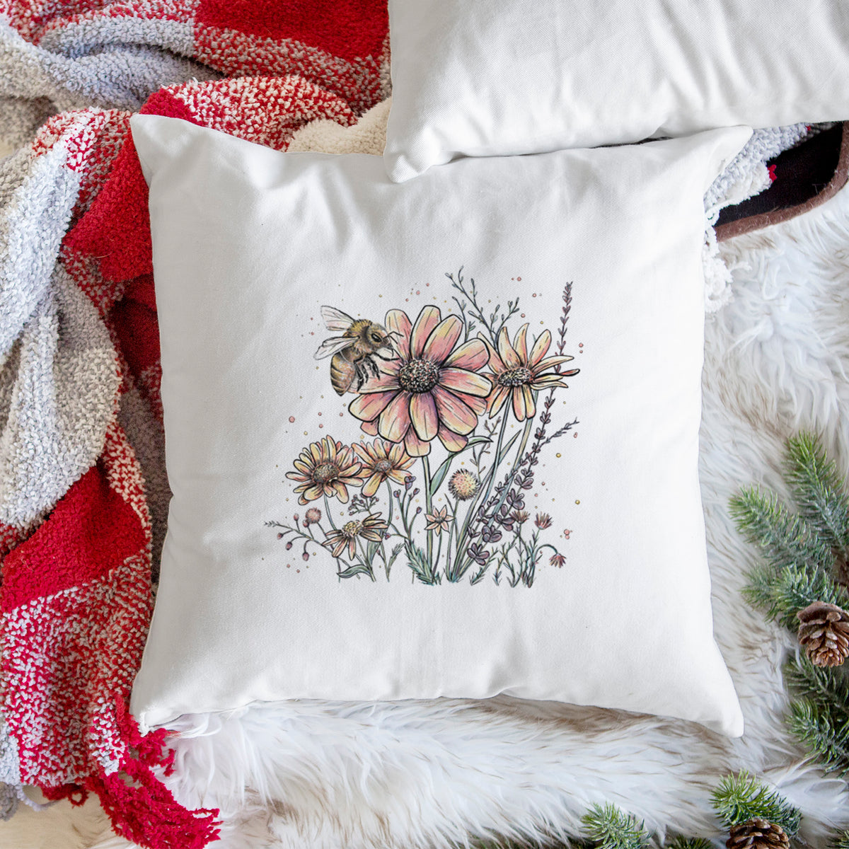 White pillow cover printed with a soft full-color illustration of a bee flying over pink and purple wildflowers, on a woven chair.