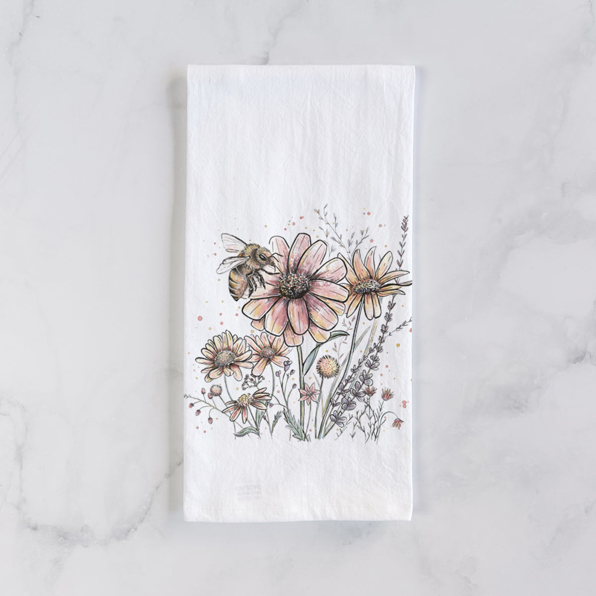 White cotton tea towel laid flat on marble, printed with a soft full-color illustration of a bee flying over pink and purple wildflowers.