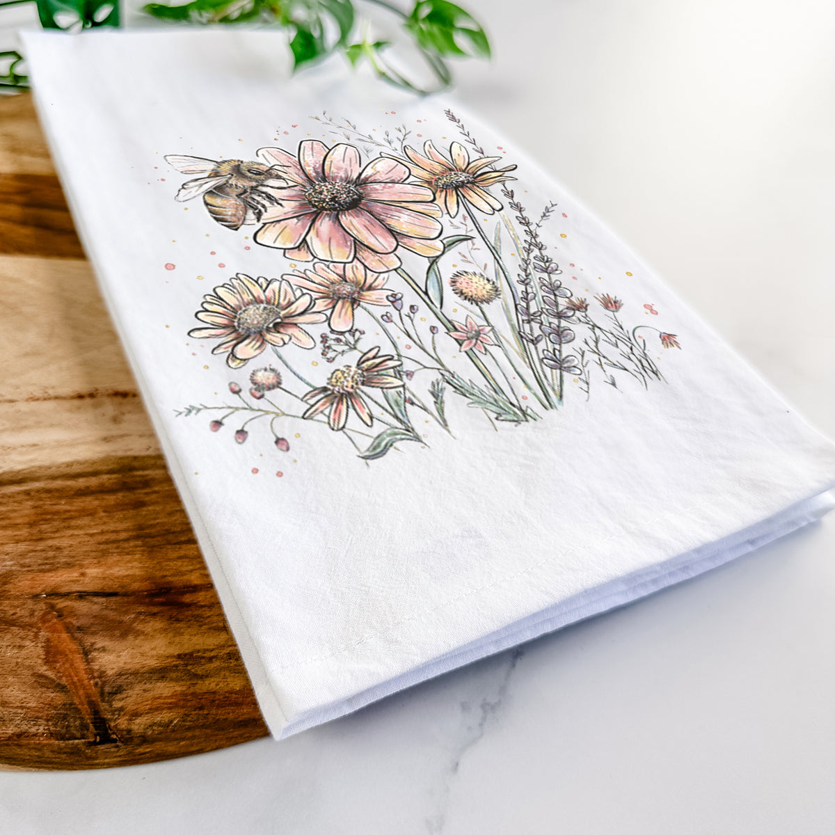 Close-up of a folded white tea towel on a wooden board, showing a soft full-color illustration of a bee flying over pink and purple wildflowers.