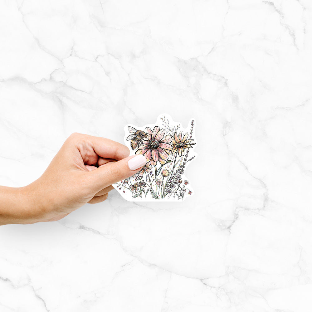 Hand holding a 4 inch vinyl decal of a soft full-color illustration of a bee flying over pink and purple wildflowers.