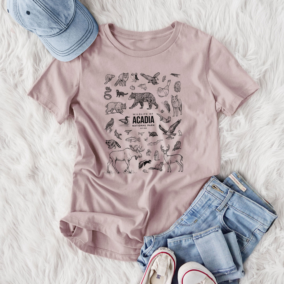 Women's relaxed fit cotton t-shirt printed with a collection of black ink wildlife illustrations surrounding the words Acadia National Park, styled flat on a white faux fur rug with jeans.