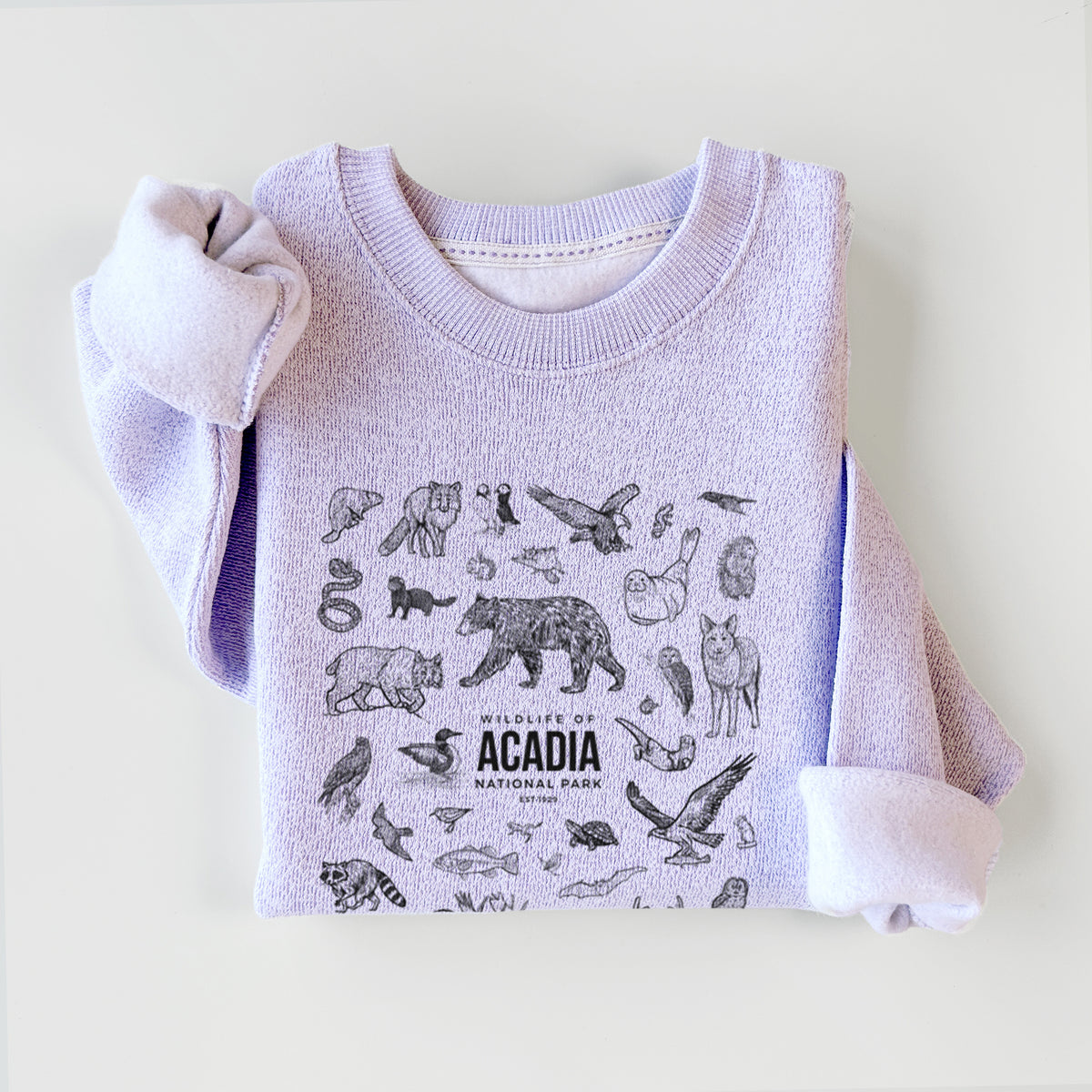 Ice purple knit sweatshirt printed with a collection of black ink wildlife illustrations surrounding the words Acadia National Park, styled flat.