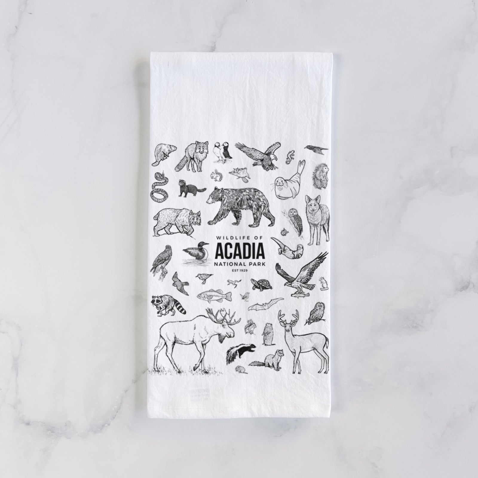 White cotton tea towel printed with a collection of black ink wildlife illustrations surrounding the words Acadia National Park, on a marble counter beside a round wooden cutting board and greenery.