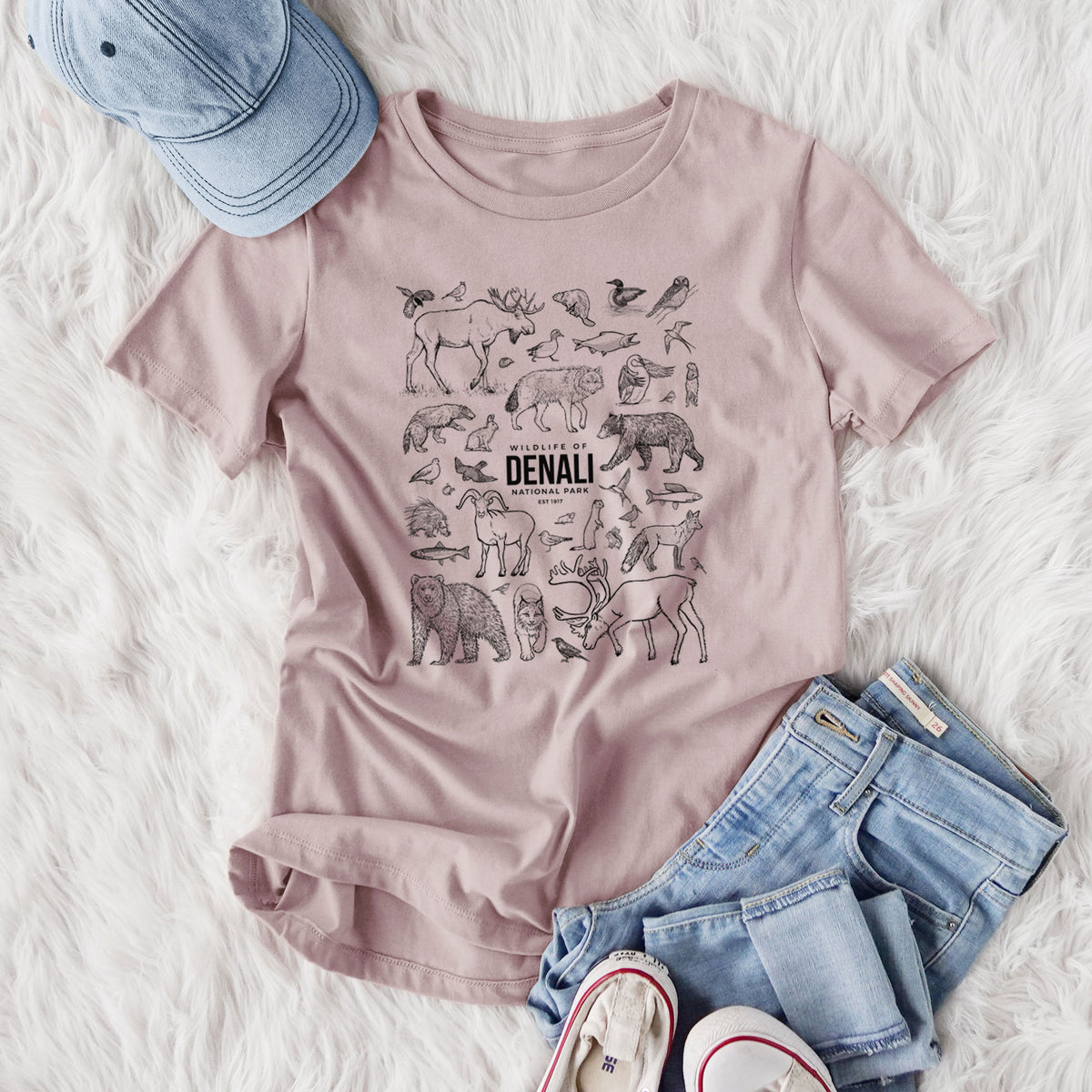 Women's relaxed fit cotton t-shirt printed with a collection of black ink wildlife illustrations surrounding the words Denali National Park, styled flat on a white faux fur rug with jeans.
