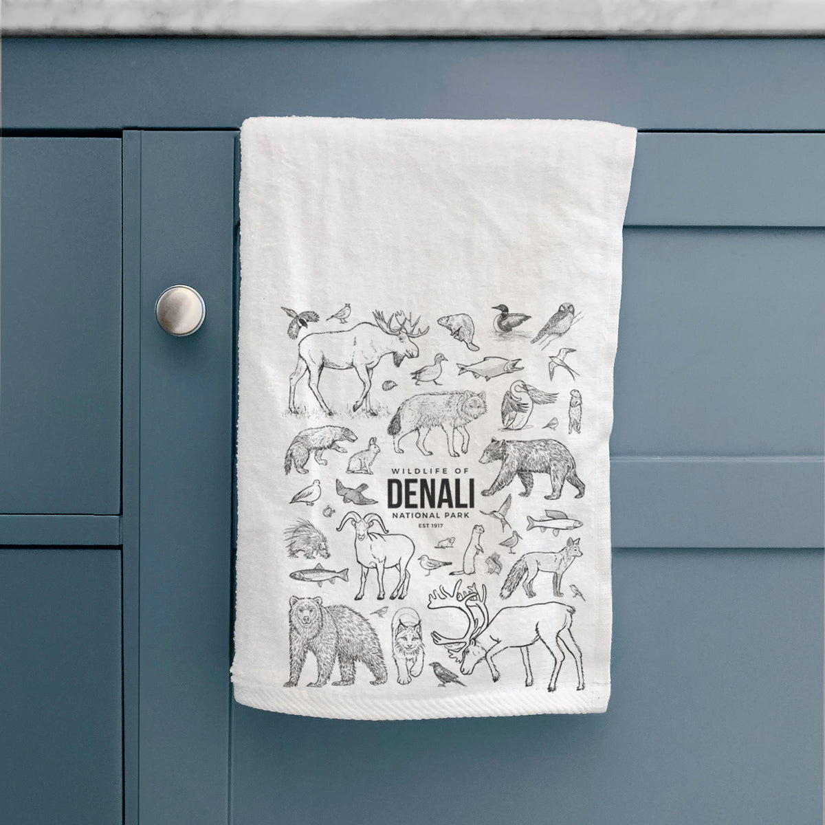 White hand towel printed with a collection of black ink wildlife illustrations surrounding the words Denali National Park, hanging on a cabinet door.