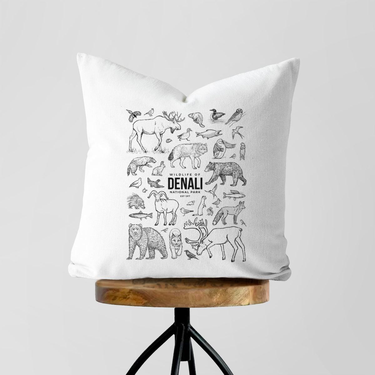 White pillow cover printed with a collection of black ink wildlife illustrations surrounding the words Denali National Park, on a wooden stool.